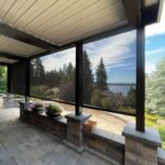A covered patio with gray stone tiles and partial black mesh screens provides ample shade as it overlooks a scenic view of trees, a distant body of water, and a partly cloudy sky. Potted flowers and plants decorate the stone ledge.