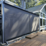 A modern patio with dark retractable outdoor screens providing shade on one side, attached to a house with large windows and surrounded by trees. The deck features brown wooden planks, small steps, and a sleek pergola overhead.