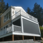 A two-story house with beige siding and white trim features a large screened-in porch on the second floor, offering relaxing shade beneath its white railings, set against a backdrop of trees and blue sky.