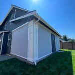 A modern house with dark siding and white trim features large exterior shade pergolas covering the windows on one side. The house sits on green grass under a clear blue sky with sunlight shining above.