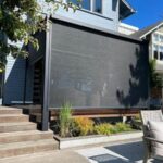 A modern house with a covered patio and pergola, featuring a large retractable black shade screen, wooden steps, outdoor seating, and landscaped plants in front of the patio area.