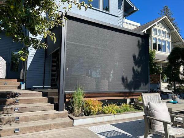 A modern house with a covered patio and pergola, featuring a large retractable black shade screen, wooden steps, outdoor seating, and landscaped plants in front of the patio area.