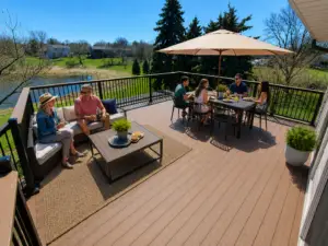 Six people enjoy a meal on a spacious outdoor deck, beautifully designed after a recent installation, complete with a table, umbrella, and lounge seating. The scene is sunny with green trees and a pond in the background.