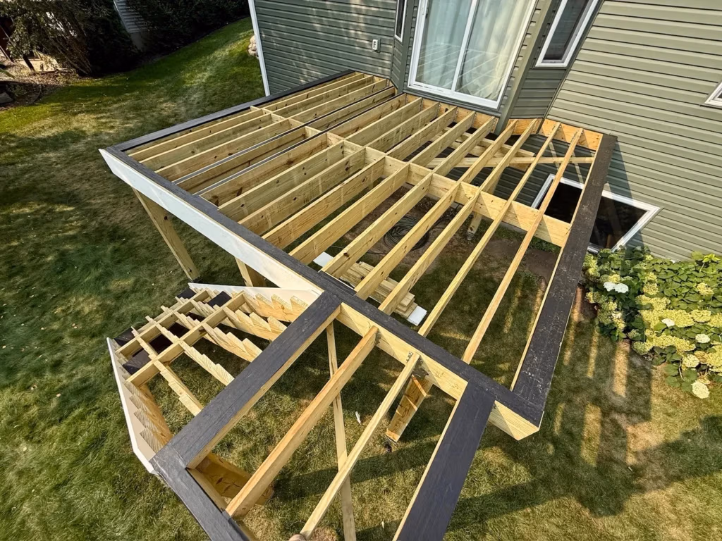 A partially constructed wooden deck is attached to a house, with exposed beams and stairs but no flooring yet. The structure sits on a grassy lawn beside green siding and a flowering bush.